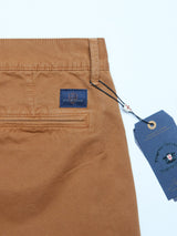 A close-up of Franco Genes Chino pants in Biscotto by Blue de Gênes shows a rear pocket with a small blue logo tag. A dark blue hang tag with text and wax seal is tied to the garment-dyed Saia twill. The pants are displayed on a light background.