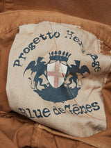 A fabric label on the brown Franco Genes Chino - Biscotto by Blue de Gênes displays a crest with a red cross flanked by two lions. The words Progetto Helv. and Blue de Gênes are printed. The garment-dyed label is slightly frayed at the edges.