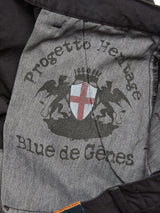 Close-up of a fabric label inside the Franco Genes Chino - Charcoal. It features the text Progetto Heritage and Blue de Gênes around a crest with a red cross on a white shield, flanked by two lion-like figures on gray, complementing the chino craftsmanship.