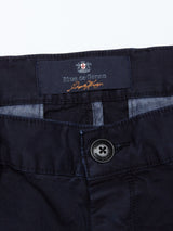 A close-up of Franco Genes Chino - Navys waistband reveals a black button and an interior label. The Blue de Gênes logo in red, white, and black is partially visible against garment-dyed Saia twill fabric.