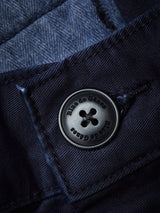 Close-up of a dark navy button on the Franco Genes Chino - Navy by Blue de Gênes, featuring the brands name engraved. The textured fabric looks like Saia twill or denim, with visible stitching around the button.