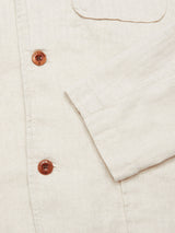 Close-up of the Gabbiano Winnot Blazer by Blue de Gênes, made from textured natural cotton linen with a herringbone pattern, brown buttons, and visible stitching. The sleeve and part of the shirts front are shown.