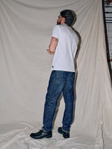 A man with short hair and a beard, facing away from the camera, wears a relaxed fit white t-shirt, Giacomo 77 Dark Used Jeans in mid-blue denim by Blue de Gênes, and black shoes. He poses confidently against a light-colored fabric backdrop.