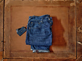 A pair of Blue de Gênes Giacomo 77 Dark Used Jeans in mid blue denim with rolled cuffs lies on a rustic wooden surface. A new tag dangles from the rigid denim, while the worn wood underneath adds to the jeans vintage vibe.