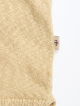 A close-up of the BGjohn Slubby Polo in Pale Khaki by Blue de Gênes, showing its beige slubby cotton knit fabric and a side tag with a red cross on a white shield. The detail stands out against the white background, highlighting vintage leisurewear style.