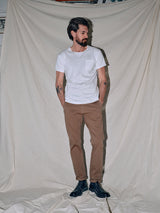 A dark-haired, bearded man stands by a beige curtain, wearing a white T-shirt and Blue de Gênes Menzo Dobby Chino in Brownie. Black shoes complete his look, and tattoos peek from his forearms. Hands in pockets, he gazes sideways, embodying relaxed comfort stretch style.
