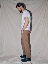 A man with a beard stands in profile against a draped fabric backdrop, exuding vintage vibes. He wears a white t-shirt, Blue de Gênes Menzo Dobby Chino - Brownie pants, and black shoes. His hands rest in his pockets as he appears deep in thought.