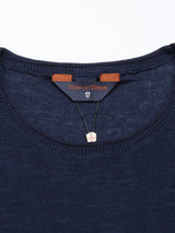 A close-up of the Monte Knit - Navy crewneck by Blue de Gênes reveals its dark blue neckline, adorned by a small necklace with a rectangular red and white cross pendant. The brown leather label on the elegant cotton/linen blend adds sophistication.