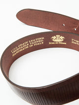 A close-up of the Blue de Gênes Obla Belt in Dark Brown reveals detailed craftsmanship. The inside displays gold text—FULL GRAIN LEATHER, VEGETABLE TANNED, HANDMADE IN ITALY—and decorative symbols that hint at an antique brass buckle and elegance.