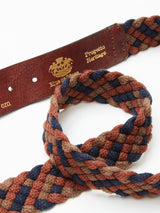 A close-up of the Pratese Belt by Blue de Gênes showcases its brown, blue, and rust woven loop. Made with braided cotton webbing, it rests next to a leather strap embossed with gold text and logos. Handmade in Italy, it reflects fine craftsmanship through intricate interlacing patterns.