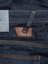 Close-up of regular slim-fit Repi Dai V Dark Jeans in dark blue denim by Blue de Gênes showcases orange stitching, brown leather patch with subtle text, belt loop, and part of a back pocket. The comfort stretch fabric has a slightly textured appearance.
