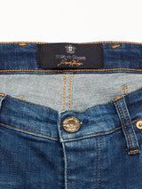 Close-up of Blue de Gênes Repi Dragon Dark Used Jeans in denim blue, featuring a button with intricate design and a waistband label. The regular slim-fit style highlights visible stitching, emphasizing the comfort stretch denim texture and detailed craftsmanship.
