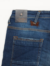 A close-up of Blue de Gênes Repi Dragon Dark Used Jeans in Denim Blue highlights the comfort stretch denim. The waistband has belt loops, yellow stitching on edges, a leather brand patch near the pocket, and a black tag attached with a metal pin.