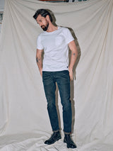 A bearded man in a white T-shirt and jeans stands before a light backdrop. Wearing Repi Haworth Used Jeans by Blue de Gênes, he looks down with a slight smile, revealing arm tattoos. Hes sporting black shoes.