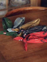 On a wooden table lies a vibrant collection of cotton bandanas in green, blue, tan, black, and Red by Blue de Gênes. Each features artistic designs with tied ends, with some showcasing a faded logo print for added character.