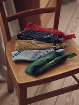 A wooden chair holds four Blue de Gênes Bandana - Celeste cotton bandanas in red, black, blue, and green, each with a unique pattern for a subtle fashion statement. The chair sits gracefully on a light wooden floor.