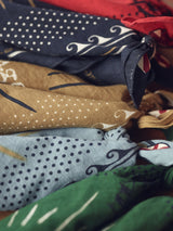 A close-up of the Bandana - Celeste by Blue de Gênes displays vibrant, patterned cotton fabrics tied into knots. The scarves, in blue, green, brown, and red hues with dots and abstract designs, make a bold fashion statement.