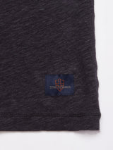 A close-up of a dark, natural texture fabric shows a small rectangular patch featuring an orange shield emblem with the words since 7 genes on a black background, located near the edge of what appears to be part of a Totti T-Shirt in Dark Navy from Blue de Gênes.