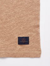 A close-up shows the beige, textured linen jersey of the Totti T-Shirt - Tuape by Blue de Gênes, featuring a rectangular dark blue patch with an orange shield design and unreadable text logo. The natural textures edge displays a meticulously hemmed finish.