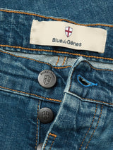 Close-up of Vinci Topaz Vintage Jeans - Used by Blue de Gênes in blue comfort stretch denim, featuring exposed branded buttons, a label with a red cross above the brand name, orange stitching, and a blue buttonhole.