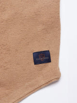 Close-up of beige knit fabric from a Zorba Knit Waistcoat – Taupe by Blue de Gênes, featuring a dark blue label with a stylized emblem and text Nudie Jeans in orange. The merino wool displays a subtle textured pattern.