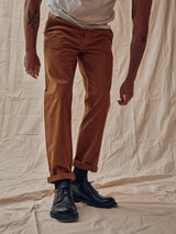 A person wearing Blue de Gênes Franco Genes Chino in Biscotto, a white shirt, and black shoes stands against a wrinkled beige fabric backdrop. Tattoos on their arms blend seamlessly with their relaxed posture, showcasing the outfits casual elegance.