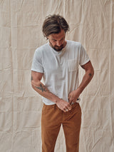 A tattooed man in a Blue de Gênes Sagi Nuovo T-shirt, made of premium cotton, adjusts his regular-fit brown pants. Against a beige crumpled fabric backdrop, he looks down at his hands, highlighting the refined details of his outfit.