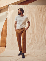 A man with a beard stands against a beige backdrop, showcasing effortless style in a white t-shirt and Blue de Gênes Franco Genes Chino in Biscotto, paired with black boots. With one hand in his pocket and the other by his side, he gazes thoughtfully to his right.