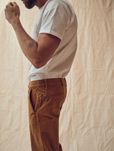 A man in a white t-shirt and Franco Genes Chino - Biscotto by Blue de Gênes stands sideways against a beige backdrop. His face is hidden, with a raised left arm revealing a small forearm tattoo. The image highlights the garment-dyed texture of his clothing and relaxed posture.