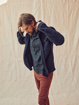 A man with medium hair wears a dark blue jacket over a button-up shirt and Blue de Gênes Paulo Tucan Chino pants in Chipotle. He stands against a beige fabric backdrop, gazing downward.