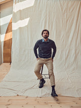 A man with a short beard and medium-length hair sits on a stool against draped white cotton fabric. He wears a dark sweater over the Blue de Gênes Miguel Chambray Shirt - Lt. Denim Blue, beige pants, and black boots, in a room with wooden floors and soft natural light.