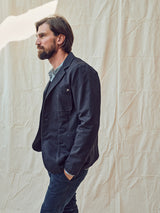 A bearded man dressed in a dark jacket and Paulo Tucan Chino - Navy by Blue de Gênes stands in profile against a white fabric backdrop.