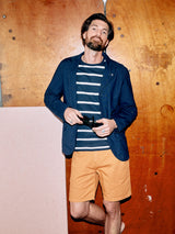 A bearded man leans against a wooden wall, exuding an artistic vibe. He wears a navy blazer over a garment-dyed striped shirt and Menzino Shorts in apricot by Blue de Gênes, holding sunglasses.