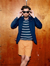 A bearded man poses against a wooden background, adjusting his sunglasses. He wears a blue blazer, a navy and white striped shirt, and Blue de Gênes Menzino Shorts in Apricot. His messy hair completes the relaxed yet stylish look.