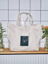 The BDG Canvas Bag - Off White by Blue de Gênes, featuring a blue patch with text, hangs on a wooden shelf for everyday use. Two glass vases with orange and yellow flowers are beautifully arranged on either side against a white tiled wall.