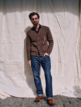 A bearded man wearing glasses sports a relaxed loose fit, donning a brown cardigan with embroidered design, navy shirt, Giacomo 77 Dark Used Jeans by Blue de Gênes in mid-blue denim, and brown shoes. He stands on cobblestones against a wrinkled white cloth backdrop.