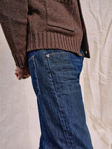 A person in a brown sweater and Giacomo 77 Dark Used Jeans by Blue de Gênes stands against a neutral, textured backdrop. The side view highlights the relaxed loose fit of the clothing, emphasizing its stylish textures and colors.
