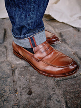 A close-up captures someone wearing brown leather loafers with cuffed Giacomo 77 Dark Used Jeans in mid blue denim by Blue de Gênes. They stand on cobblestone pavement, where black socks with red stripes and lighting enhance the shoe texture and stone surface.