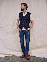 A bearded man stands against an off-white fabric, wearing a Blue de GĂȘnes Zorba Knit Waistcoat in navy over a white shirt, with blue jeans and brown shoes. His hands are in his pockets, admiring the corozo nut buttons as he gazes sideways.