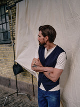 A brown-haired man in a white shirt and Blue de GĂȘnes Zorba Knit Waistcoat - Navy stands with arms crossed before a cream fabric backdrop, gazing sideways by a brick wall and window with corozo nut buttons decor.