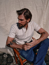 A tattooed man with a beard sits on a wooden chair against a neutral backdrop, wearing the Nipote AW S/S Grandad - Whitecap from Blue de Gênes in cotton interlock jersey and blue jeans. In the sunny outdoor setting, he holds a cigarette and gazes sideways.