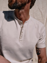 A bearded person wears a Blue de Gênes Nipote AW S/S Grandad T-Shirt in Whitecap, sitting against a textured beige backdrop. A tattoo is partly visible on their left arm, while the cotton interlock jersey fabric casts soft shadows, highlighting the shirts classic design.