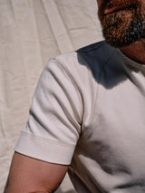 Close-up of an individual wearing a Blue de Gênes Nipote AW S/S Grandad in Whitecap. The focus is on the cotton interlock jerseys texture and stitching. The backdrop features a soft, neutral fabric, with just part of the persons face and arm shown.