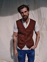 A man with brown hair and a beard stands against a beige backdrop, wearing a Blue de Gênes Zorba Knit Waistcoat - Prune over a white shirt and blue jeans. Featuring corozo nut buttons, his outfit complements his arm tattoos as he looks directly at the camera with one hand in his pocket.