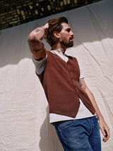 A tattooed man with a beard wears the Blue de Gênes Zorba Knit Waistcoat in Prune over a white shirt and blue jeans, looking sideways with his hand on his head. In the background, theres a light cloth backdrop and a brick archway.