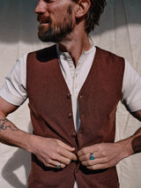 A tattooed man wearing rings adjusts his Blue de Gênes Zorba Knit Waistcoat in Prune over a white shirt, set against a light fabric backdrop.