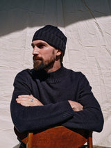 A bearded man in a dark lambswool cashmere-blend sweater and an Oscar Croft Beanie in Dark Navy by Blue de Gênes sits with arms crossed on a wooden chair. The background is a crumpled white sheet, and hes looking to the side, partially in shadow.