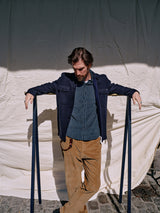 A bearded man leans against two posts on a cobblestone street, wearing a navy jacket, Blue de Gênes Enrico Cord Shirt - Blue Pattern, and baby corduroy pants. A beige cloth in the background is partially shaded by sunlight.