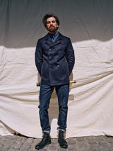 A bearded man confidently stands before a beige cloth backdrop in the Blue de Gênes Peacoat - Dark Navy, crafted from Melton wool and adorned with branded buffalo horn buttons. Paired with blue jeans and black boots, he holds a brass telescope behind his back on a cobblestone surface.