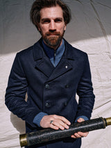 A bearded man wears a Blue de Gênes Peacoat - Dark Navy with branded buffalo horn buttons and a blue sweater, holding a vintage spyglass against a light backdrop, exuding distinguished allure.